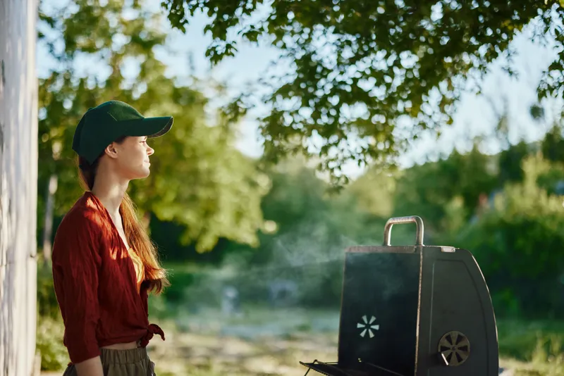 Young woman enjoying a peaceful summer day while grilling outdoors, wearing a casual outfit and a baseball cap, surrounded by lush greenery and sunlight