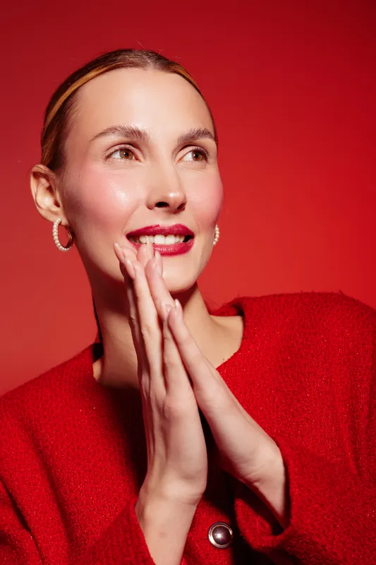 Smiling woman in red sweater with clasped hands looks away on red background. Portrait of happy female model with natural makeup and hoop earrings. Concept of joy and fashion.