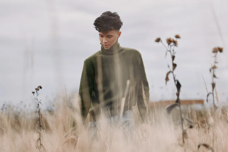 Contemplative young man in a textured green sweater walks through tall dry grass, captured in a natural candid moment that emphasizes genuine presence and understated realism.