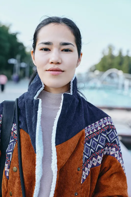 Woman wearing a colorful jacket outdoors during daytime, showcasing fashion and street style with a confident, calm mood in a sunlit park setting, highlighting texture and urban vibe