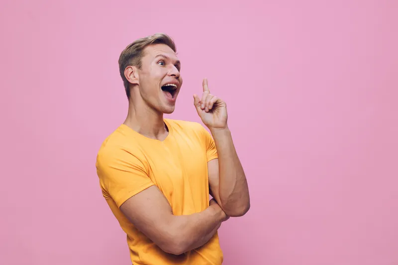 Smiling young man wearing bright yellow t-shirt expressing excitement and happiness while pointing up with finger against pink background. Studio portrait. People lifestyle concept.