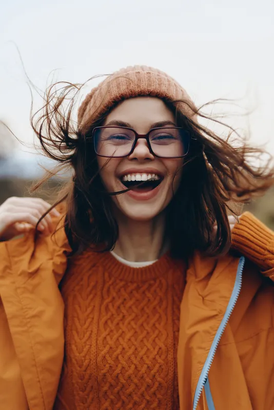 woman smile, hat and glasses, orange coat in wind, hair flying outdoors, lifestyle portrait in autumn light, casual street fashion, soft natural light
