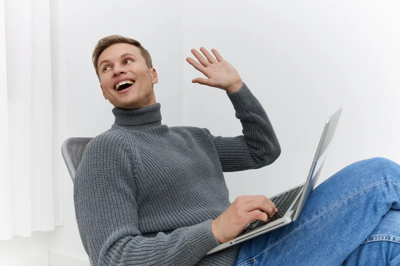 Smiling young man with short brown hair wearing casual gray turtleneck sweater and jeans, sitting with a laptop on his lap, waving happily at the camera, in a bright modern home interior, cheerful lif