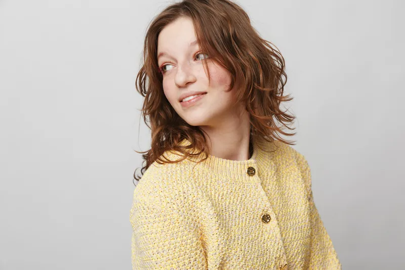 Young woman with curly brown hair wearing a yellow knitted sweater looks sideways with a gentle smile. Studio portrait on a light grey background. Concept of casual style and natural beauty.