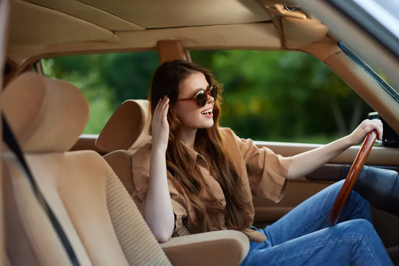 Young woman with long hair wearing sunglasses in a stylish car, enjoying a sunny day outdoors, exuding joy and confidence.