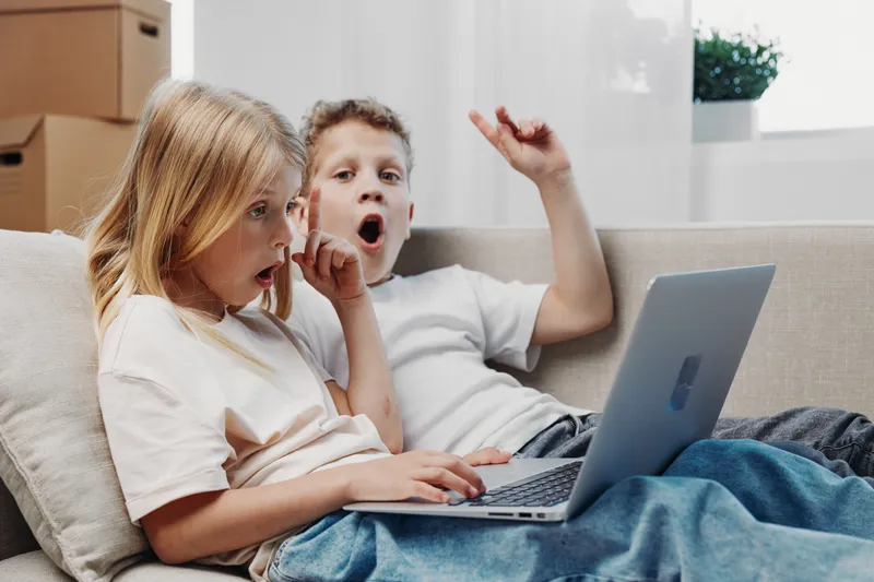 Two surprised children sitting on a sofa using a laptop together in a bright living room. Concept of technology, childhood, learning, and digital entertainment.