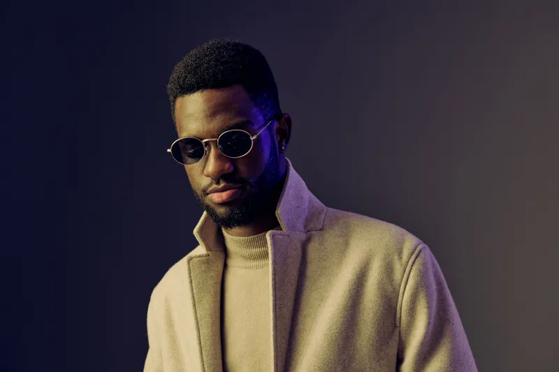 Stylish portrait of a confident Black male model wearing a fashionable beige coat and sunglasses against a dark gradient background, embodying a modern, chic vibe