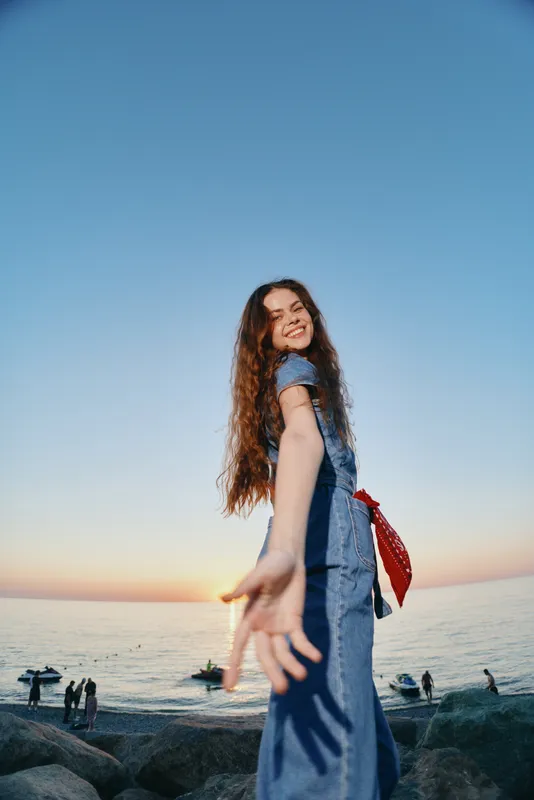 lifestyle woman in modern boho-western denim street style reaching out smiling with film color at sunset by the sea with rocks and people on the beach