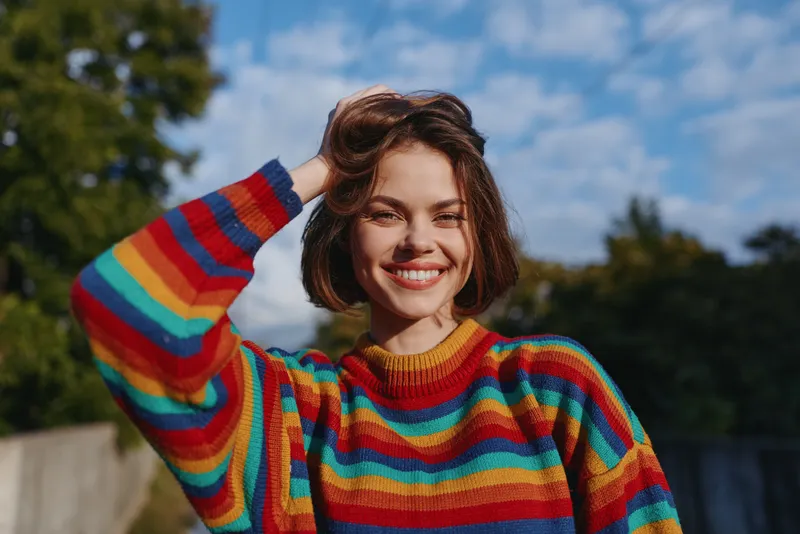 Woman smile rainbow sweater portrait outdoors happy young short hair under sunny blue sky, cheerful female in colorful knit, natural light and casual pose for lifestyle portrait.