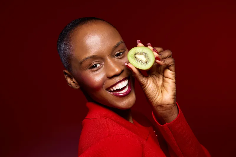 Smiling dark skinned African woman with short hair holds half a kiwi close to her face. The red background and red sweater create a vibrant contrast, concept of healthy lifestyle and fresh fruit.