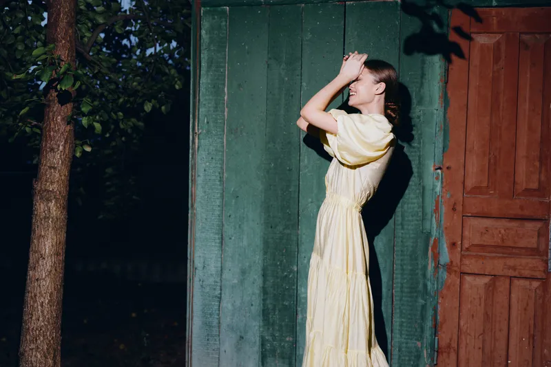 Woman in yellow vintage dress smiling in sunlight by wooden wall, authentic inclusive portrait