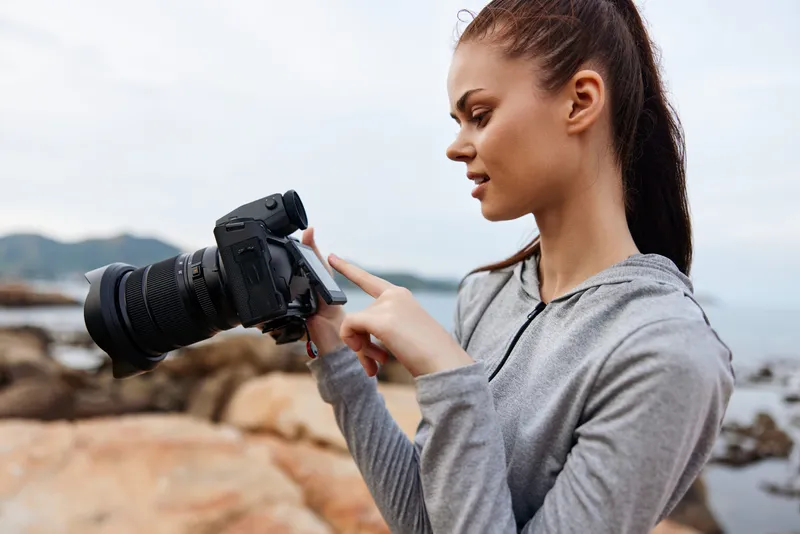 Young woman checking camera settings by the sea in a gray hoodie, displaying focus and concentration, perfect for photography enthusiasts and coastal adventure themes