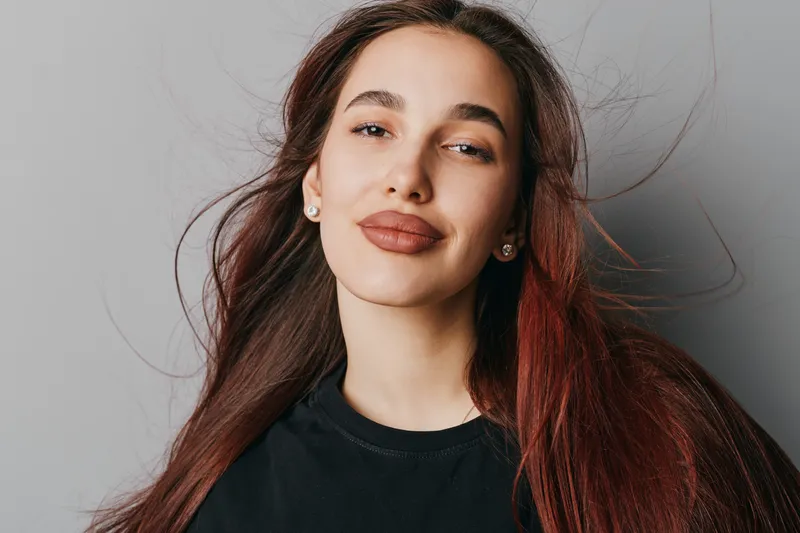 Portrait of confident woman with long brown hair and natural makeup smiling gently against neutral background. Casual style with black top and relaxed expression in studio lighting.