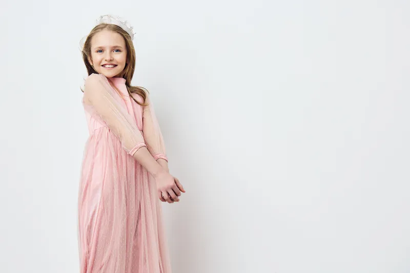 Smiling girl in a pink dress with sheer sleeves, posing against a simple white background, evoking joy and innocence, perfect for children s fashion and celebration themes
