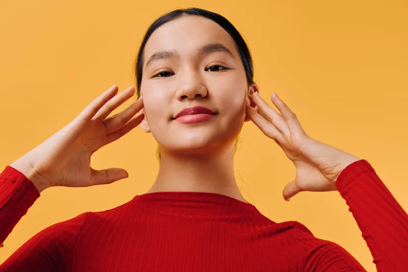 Young woman in red sweater with calm expression posing on orange background, hands near face, portrait of confident female with natural makeup and relaxed mood in studio.