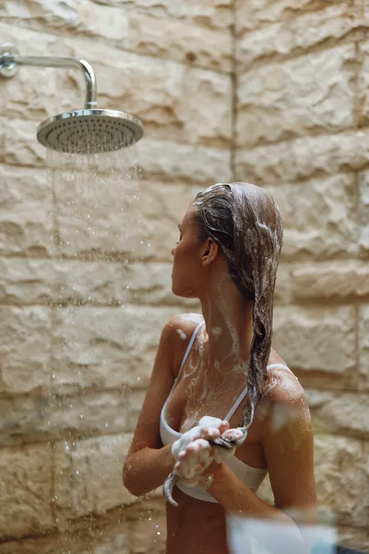 Young woman washing her long hair under warm shower in a cozy bathroom, natural stone wall background, relaxing daily hygiene routine, wet skin