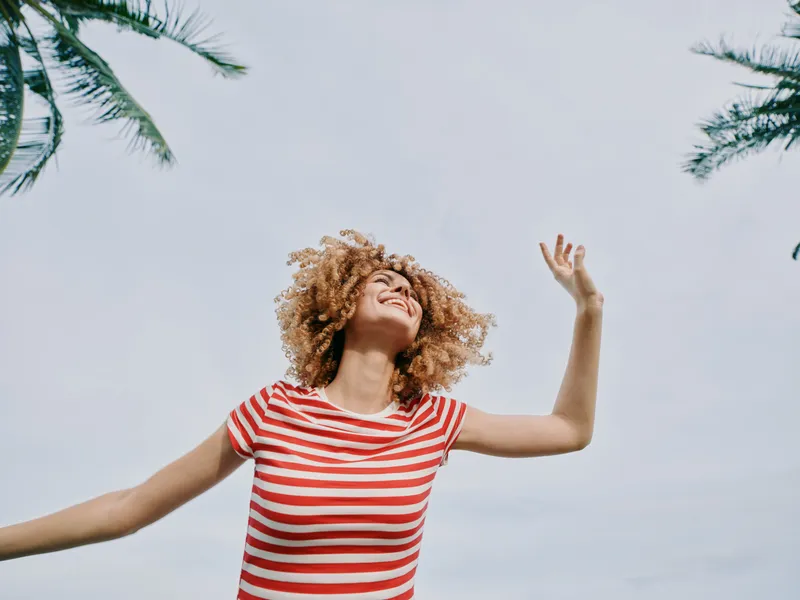 Joyful woman with curly hair in a red striped shirt raises arms toward a bright sky, capturing carefree energy, freedom, and vibrant summer mood.