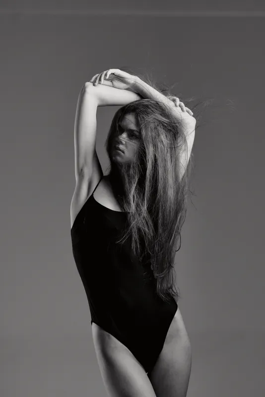 Black and white portrait of a woman with long hair posing in a black bodysuit, expressing elegance and strength.