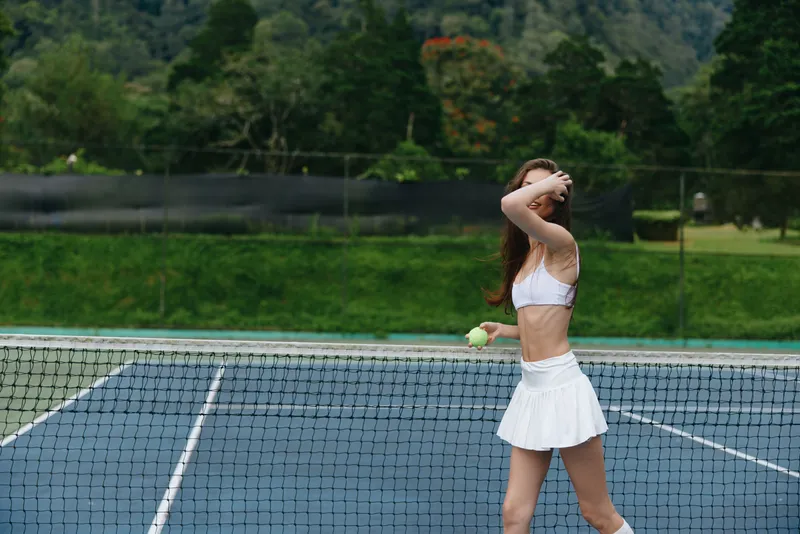 Athletic young woman playing tennis outdoors, wearing a white skirt and sports bra, displaying determination and focus against a lush green backdrop