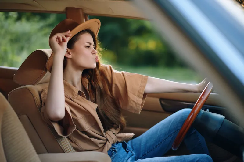 Stylish young woman driving a vintage car with a carefree expression, wearing a tan shirt and brown hat, surrounded by lush greenery.
