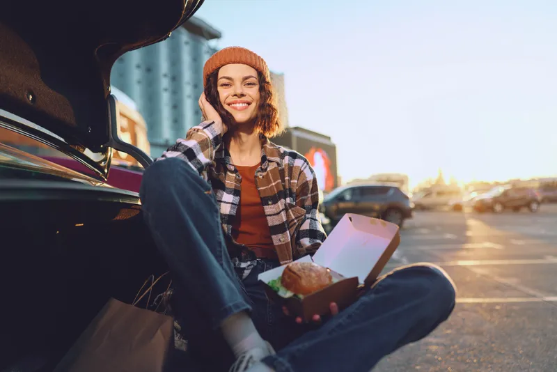 woman smile car takeout phone parking sunset casual candid lifestyle portrait of a young woman by an open trunk, talking on phone while eating takeout at golden hour glow, authenticity and mindful