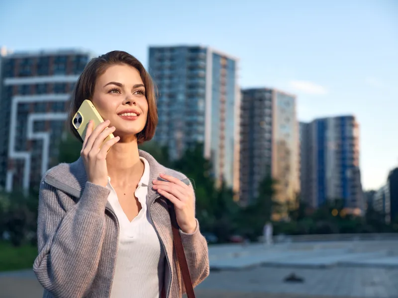 A cheerful professional woman talks on a smartphone in an urban outdoor setting, smiling and engaged, enjoying a confident moment during a busy day.