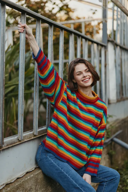 Woman smiling in a colorful striped sweater, leaning against a metal railing outdoors in an urban park, casual outfit with jeans, young adult model, cozy lifestyle portrait in golden hour.