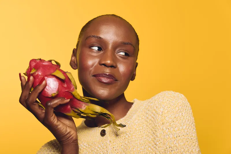 Smiling black African woman with dark skin holds dragon fruit near face, wearing yellow sweater. Studio portrait on yellow background. Concept of healthy lifestyle, fresh fruit, and natural beauty.