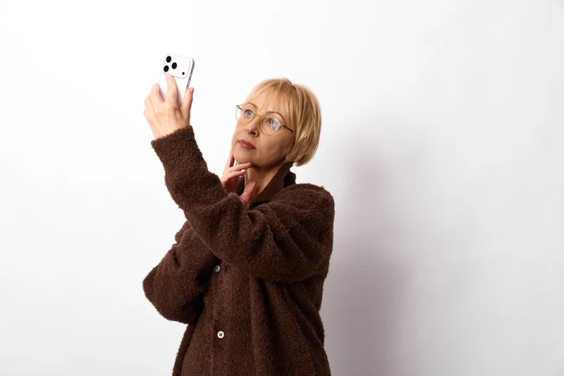 Woman with glasses holding a smartphone for communication, studio portrait on white background, focused expression, modern technology, screen light, adult lifestyle