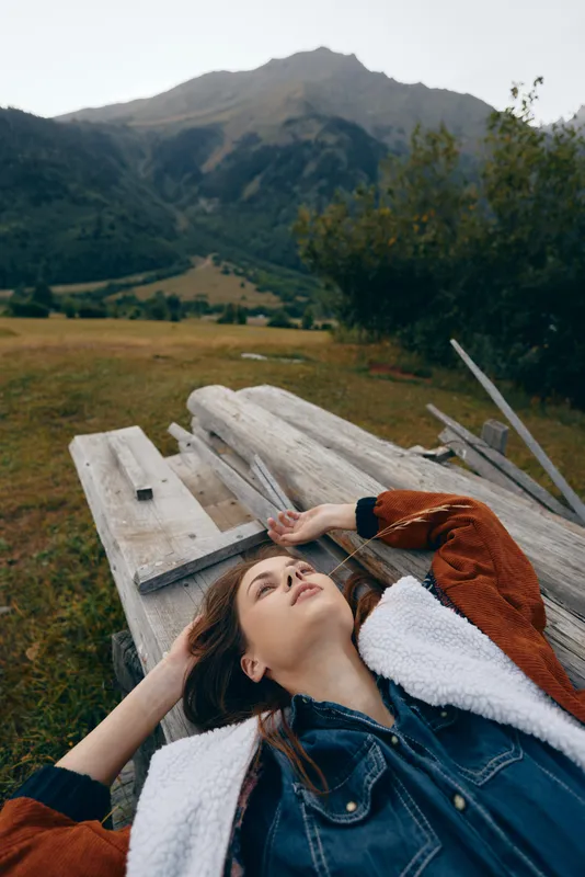 woman mountains nature relaxation bench outdoor portrait lying on wooden planks in a meadow, denim jacket and watch, peaceful mountain landscape for calm leisure and scenic escape