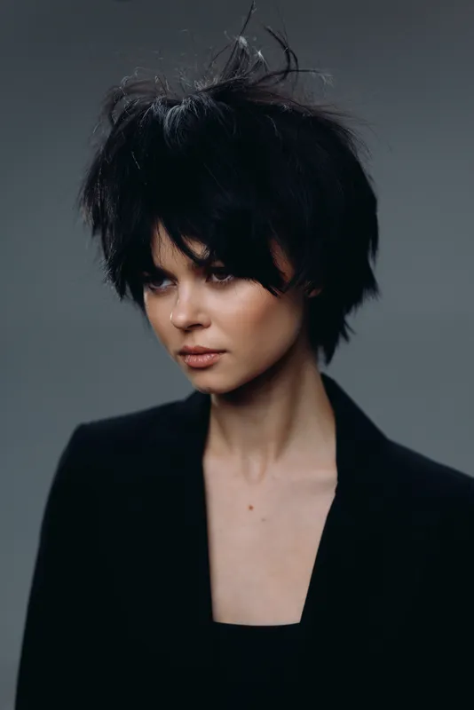 Stylish woman with short messy black hair wearing a dark blazer against a grey background, showcasing a modern and edgy fashion look.