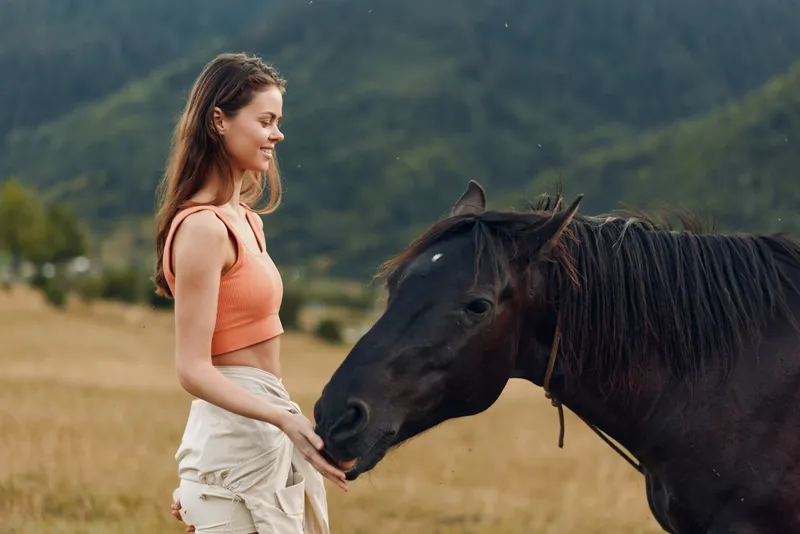 A young woman standing in a open field gently feeds a horse, offering a soft touch. She smiles warmly, sharing a calm, bonding moment with the animal.