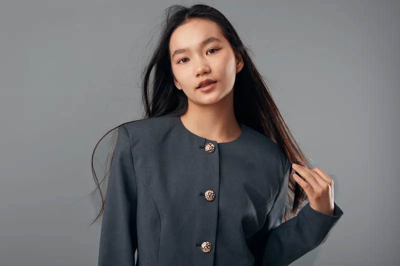 Portrait of confident young woman with long black hair wearing gray blazer with decorative buttons posing against neutral background with natural expression and calm posture.