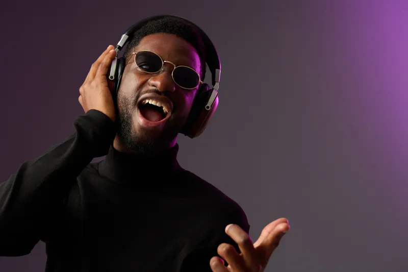 Young African American man enjoying music with headphones, wearing sunglasses, and expressing joy in a stylish, modern setting with a vibrant purple background