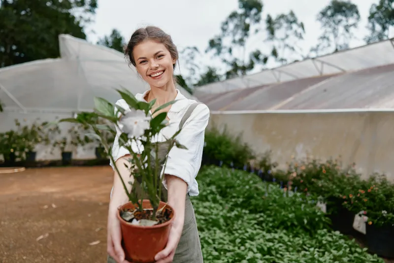 Young woman holding a potted plant in a greenhouse, smiling joyfully, surrounded by greenery, in casual outfit, early spring vibe, promoting gardening enthusiasm