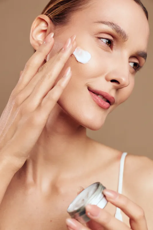 Young woman with clear skin applying white face cream on cheek. Close-up portrait with natural makeup on beige background. Concept of skincare, beauty, and self-care routine.