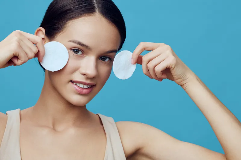 Young woman with smooth skin holding cotton pads, smiling with a fresh and natural look against a blue background, promoting skincare and self care