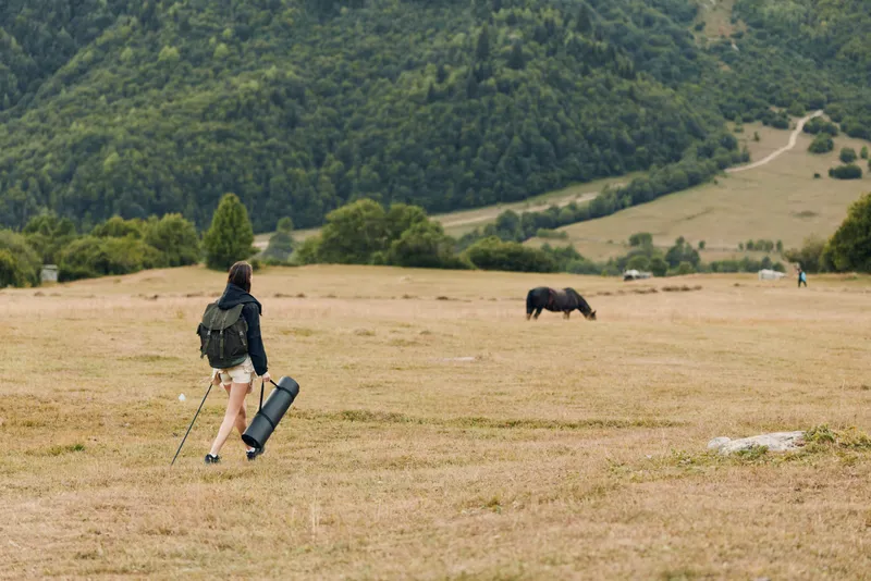 A lone traveler walks across a wide meadow with a backpack and mat, heading toward distant hills as a cow grazes nearby, creating a calm, open countryside mood.