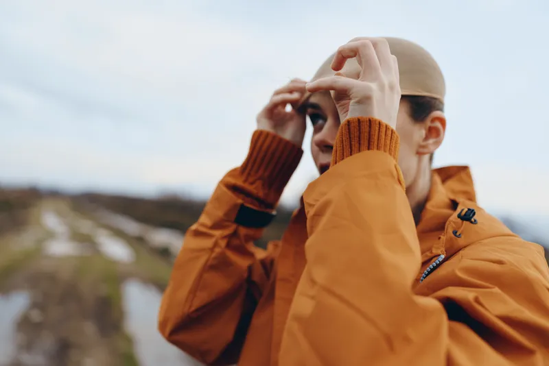 Woman lifestyle portrait in orange jacket adjusting sunglasses outdoor near mountain landscape, travel moment with soft daylight and shallow depth, focus on face and hands casually