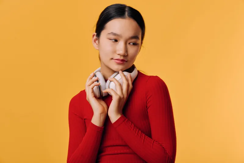 Young woman in red sweater holding headphones with hands posing on orange background looking sideways with thoughtful expression and neat hairstyle for casual style.