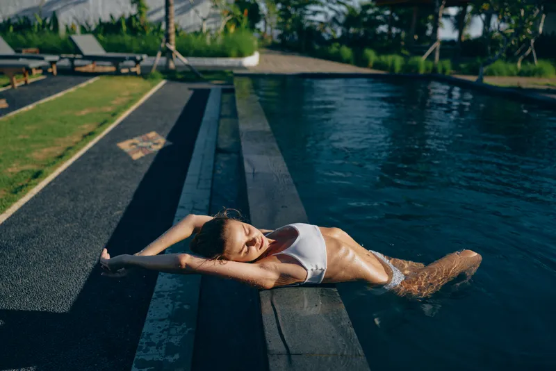 A woman rests at a pool edge, leaning on her arms, wearing a white bikini. The sunny outdoor scene conveys relaxation, wellness, and the freedom of a leisurely summer moment.