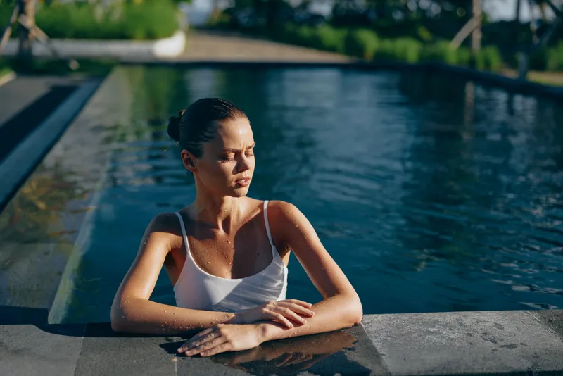 Young woman in a white swimsuit relaxes by the edge of a swimming pool with her eyes closed, enjoying a peaceful moment in the warm sunlight of a serene outdoor setting.