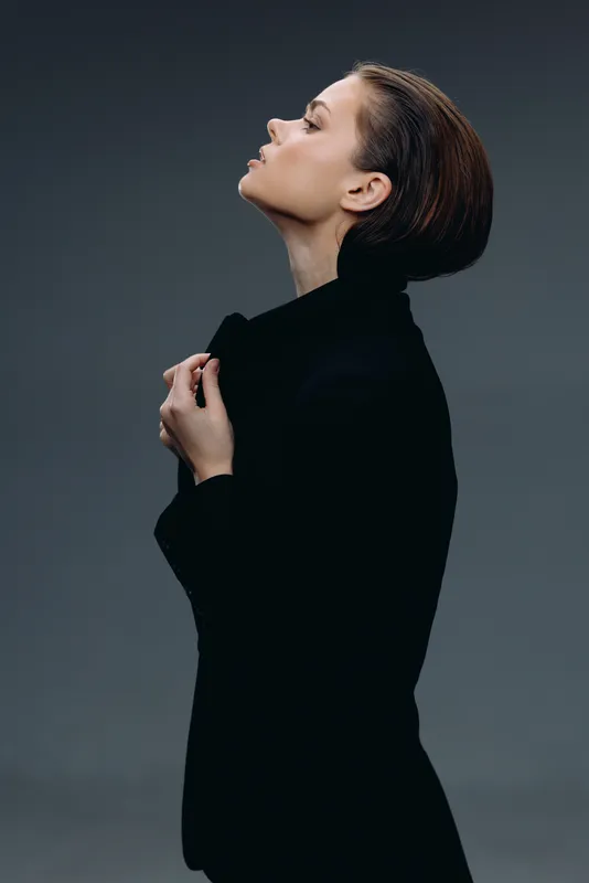 Profile of a thoughtful woman wearing a black coat against a simple gray background with soft lighting.