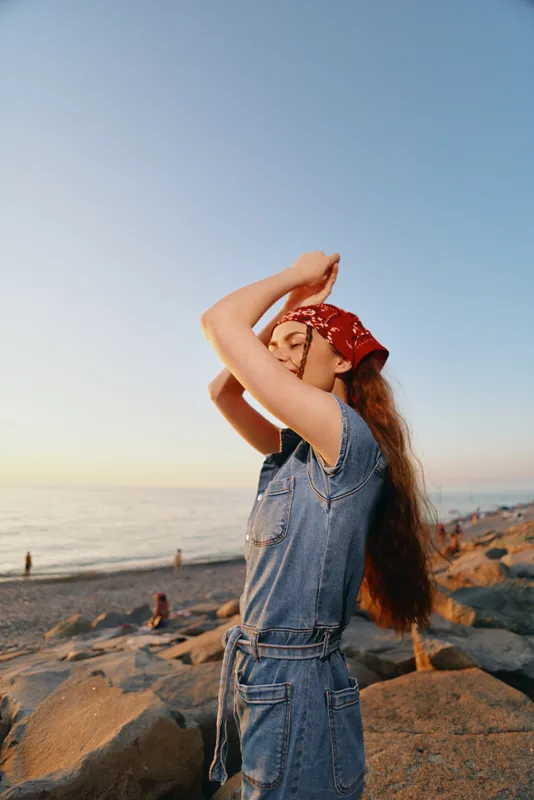 young woman wearing denim jumpsuit and red bandana enjoys calm seaside sunset with rocky shore and clear sky in warm natural light