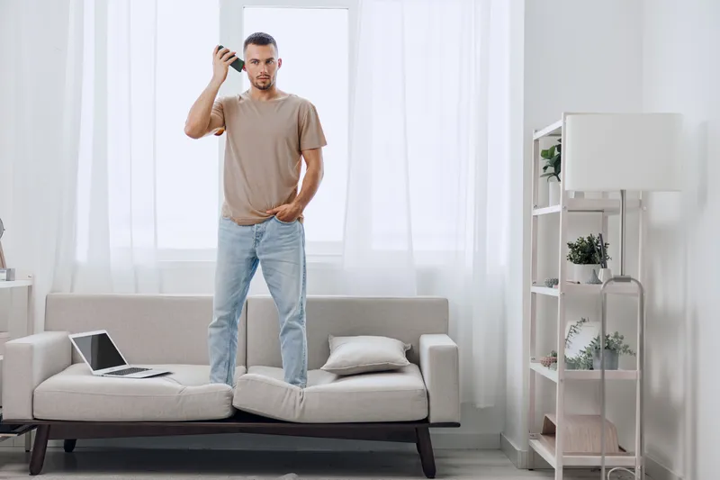 A man stands confidently on a sofa, holding a smartphone and looking thoughtfully at the screen. The bright and airy room exudes modern elegance and comfort.