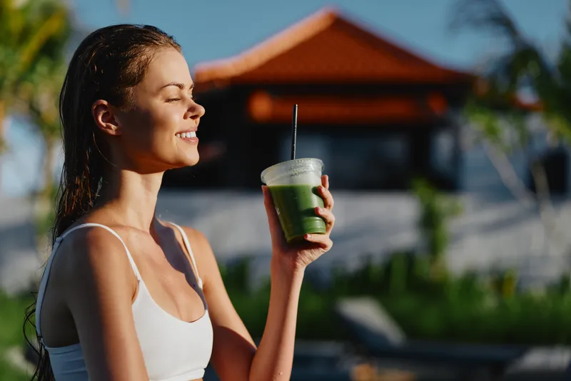 Smiling woman in a white tank top holds a green smoothie outdoors, sunlit by a pool at a tropical resort. Lifestyle, wellness, and refreshment captured in a bright, warm mood.