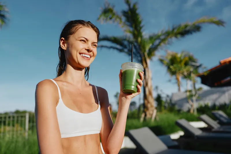 Cheerful woman outdoors, smiling under bright sun, holding a green smoothie in a clear cup, sporty white top, relaxed lifestyle moment, warm breeze, vibrant colors, palm trees in the background