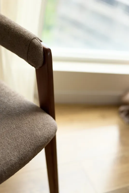 Chair armchair wood upholstery seat interior window floor in soft natural light, closeup of upholstered wooden armrest and seat near window on hardwood floor in minimalist livingroom.