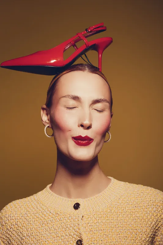 Young woman with closed eyes wears red high heel shoe on her head, dressed in yellow knit sweater. Concept of fashion, creativity, and playful style on brown background.