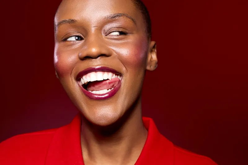 Close-up of a smiling happy black woman with dark skin and short hair wearing a red shirt. She looks to the side with bright lips and glowing skin against a red background. Joy concept.